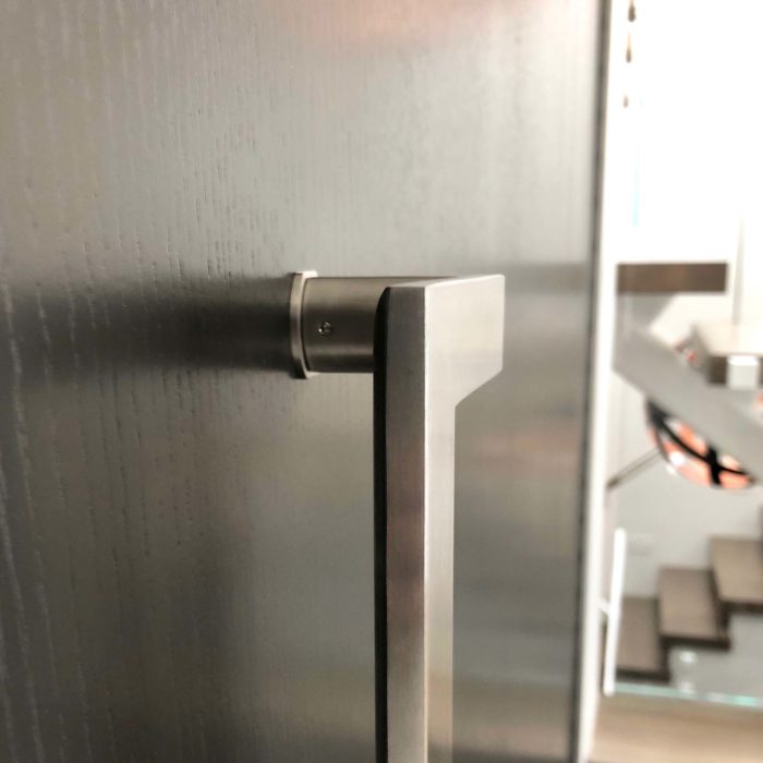 A brushed stainless steel handle mounted on a wood front door with a modern floating staircase behind.