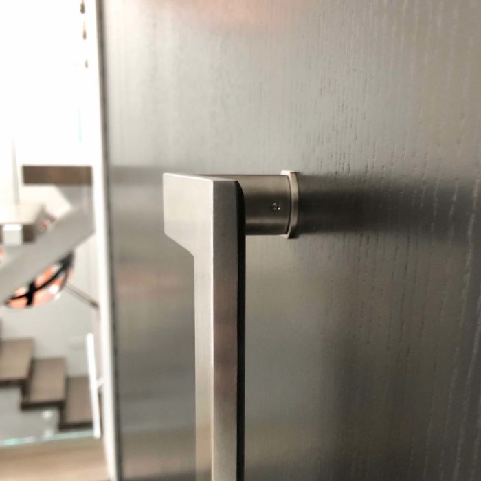 A brushed stainless steel handle mounted on a wood front door with a modern floating staircase behind.