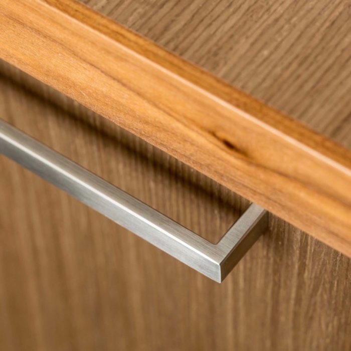 A close up view of a modern cabinet pull in brushed stainless finish mounted on a walnut cabinet.