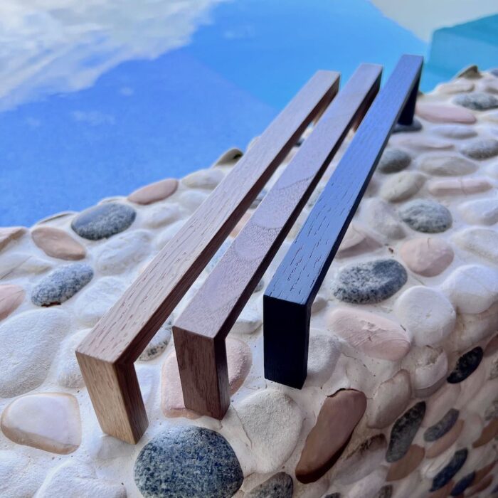 JWL Hardware's designer wooden handles in walnut, oak, and matte black finishes showcased on a pebble-strewn surface with a reflective blue water backdrop