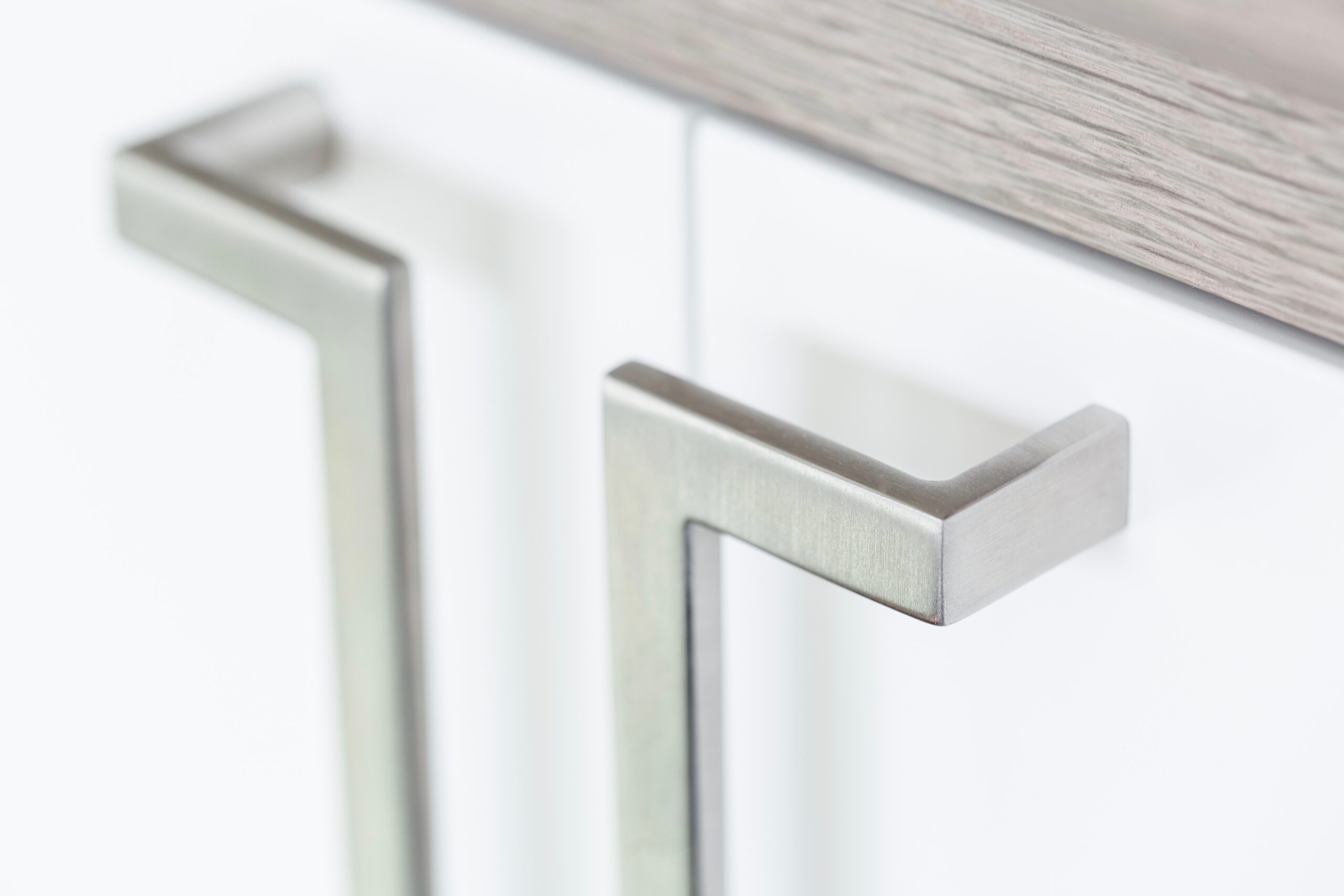 Two brushed stainless steel handles mounted on a modern cabinet with white doors and a grey oak top.