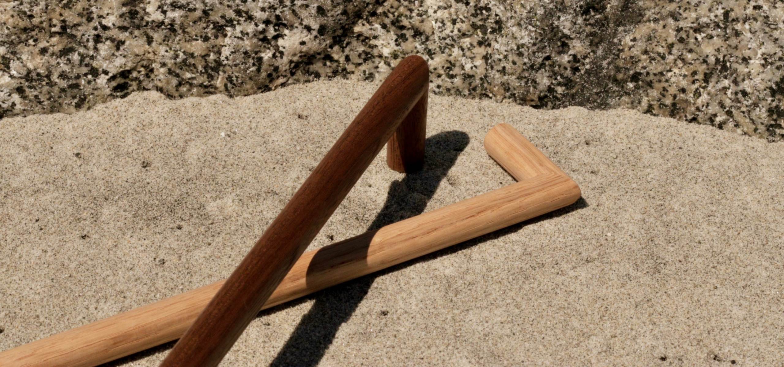 Solid wood door handles – oak and walnut Solid wood door handles in oak and walnut with a round profile, resting on sand.