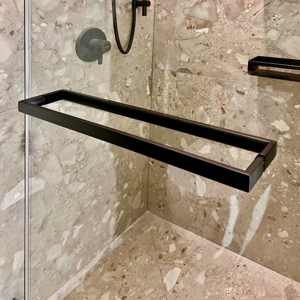 Matte black towel bar handle mounted on a frameless glass shower door, shown against terrazzo tile walls and floor